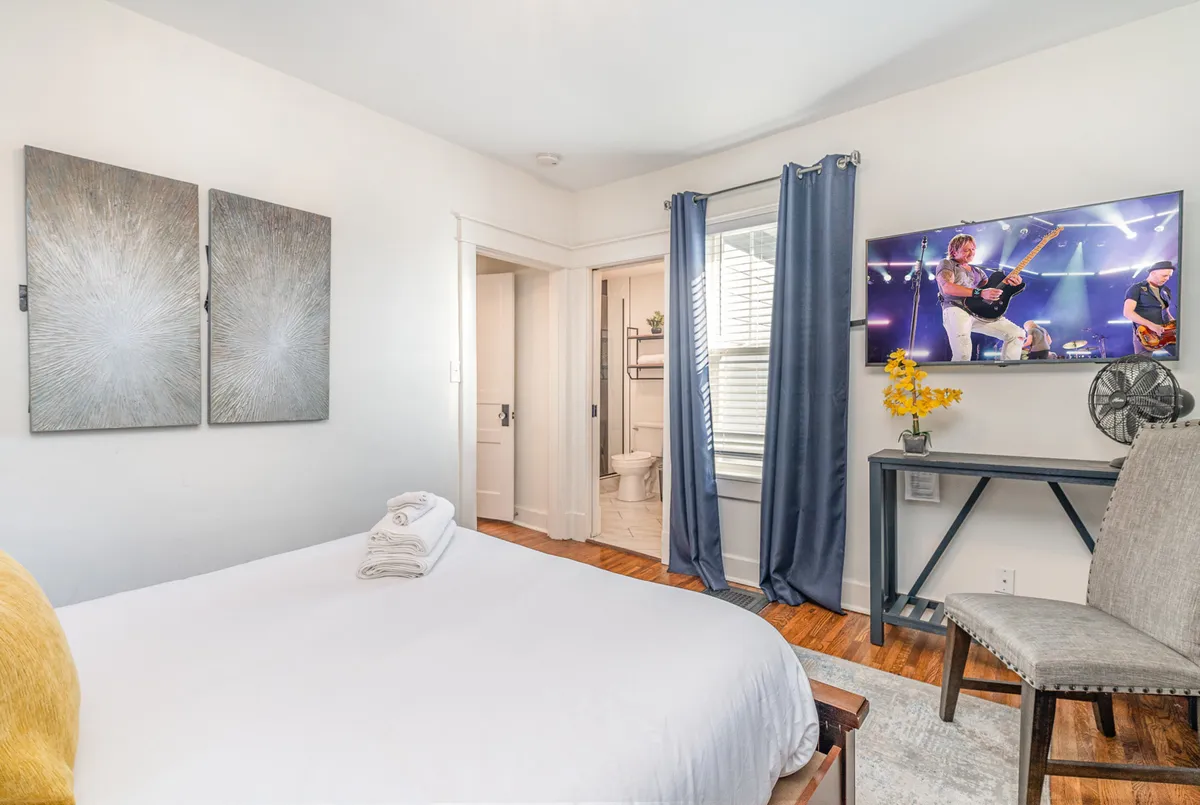 A well-appointed bedroom with white linens, a television (displaying a concert or movie), abstract wall art, blue curtains, and a small table with yellow flowers. The decor is vibrant yet relaxing, and the presence of a TV nods to the smart TV feature in the primary suite.
