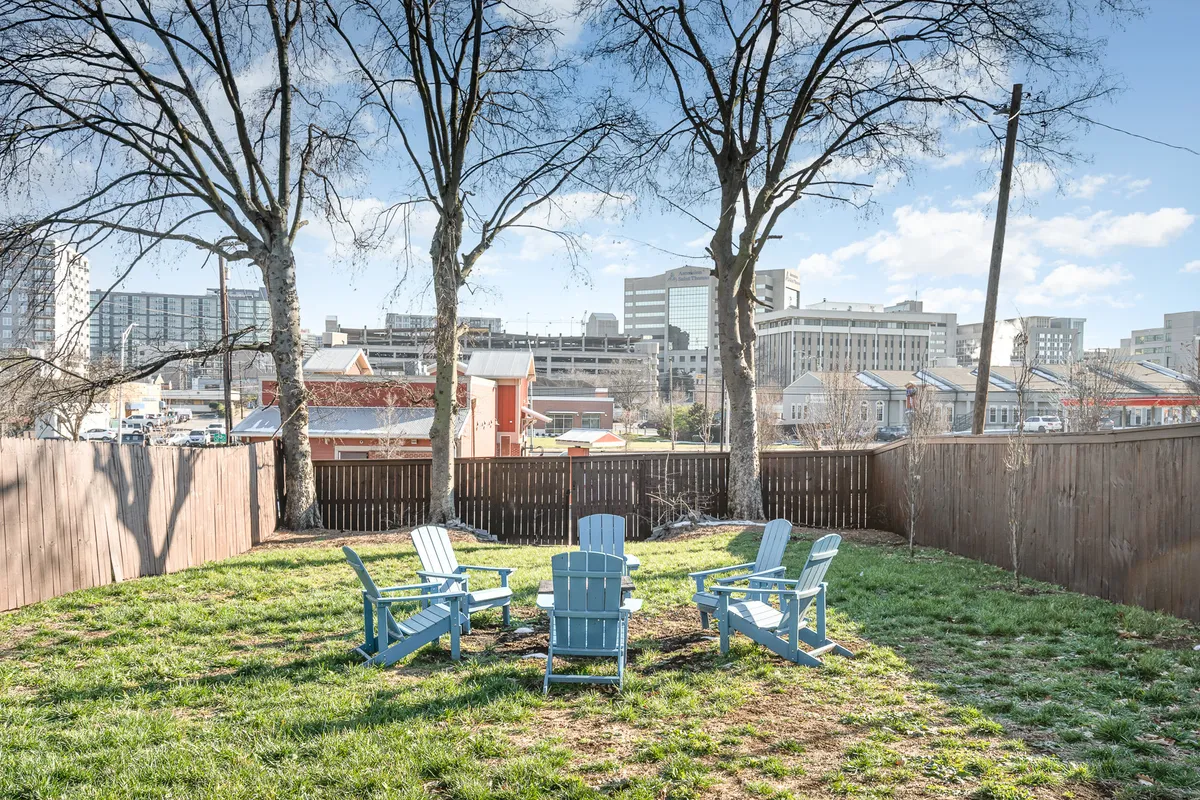 A cozy backyard scene featuring blue Adirondack chairs grouped around a fire pit, surrounded by trees and offering sunset city views. The setting is primed for outdoor gatherings, fire pit conversations, and relaxing with friends after a night out.