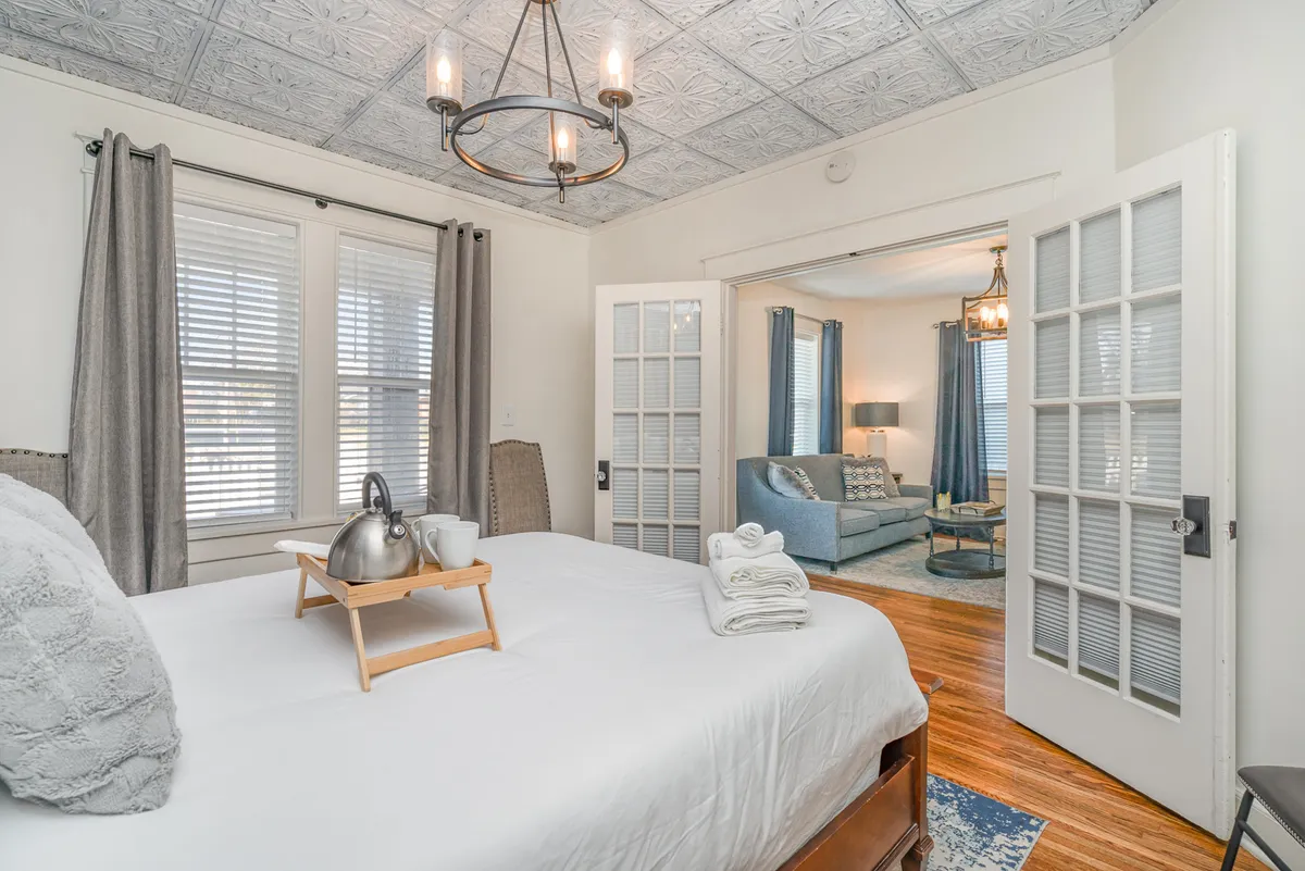 A serene bedroom with a neatly made bed, decorative pillows, a breakfast tray with teapot and cups, and a window that fills the room with natural light. The doorway leads to a connected living area, suggesting an open, homey layout.