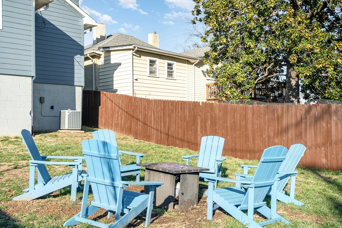 This image captures a cozy fenced backyard featuring blue Adirondack chairs arranged around a fire pit. The surrounding fence and leafy trees offer privacy and security, making it an ideal spot for gatherings or letting children play safely. The inviting setup suggests evenings spent sharing stories or roasting s'mores.