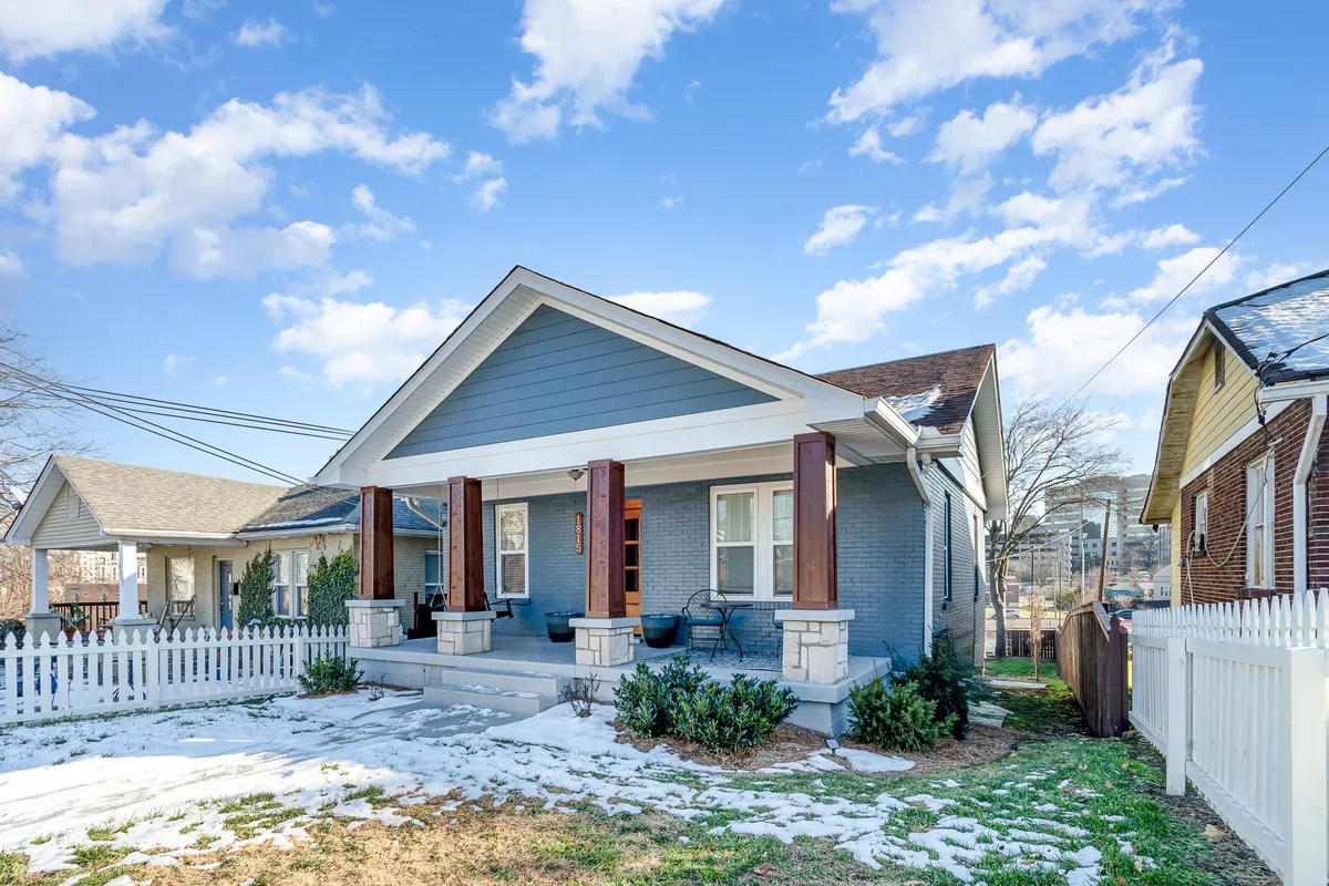 A welcoming front porch with decorative pillars and a blue-grey facade is pictured here, set against a snow-covered yard. The porch is an inviting space for relaxing, socializing, or enjoying the neighborhood view throughout the seasons.
