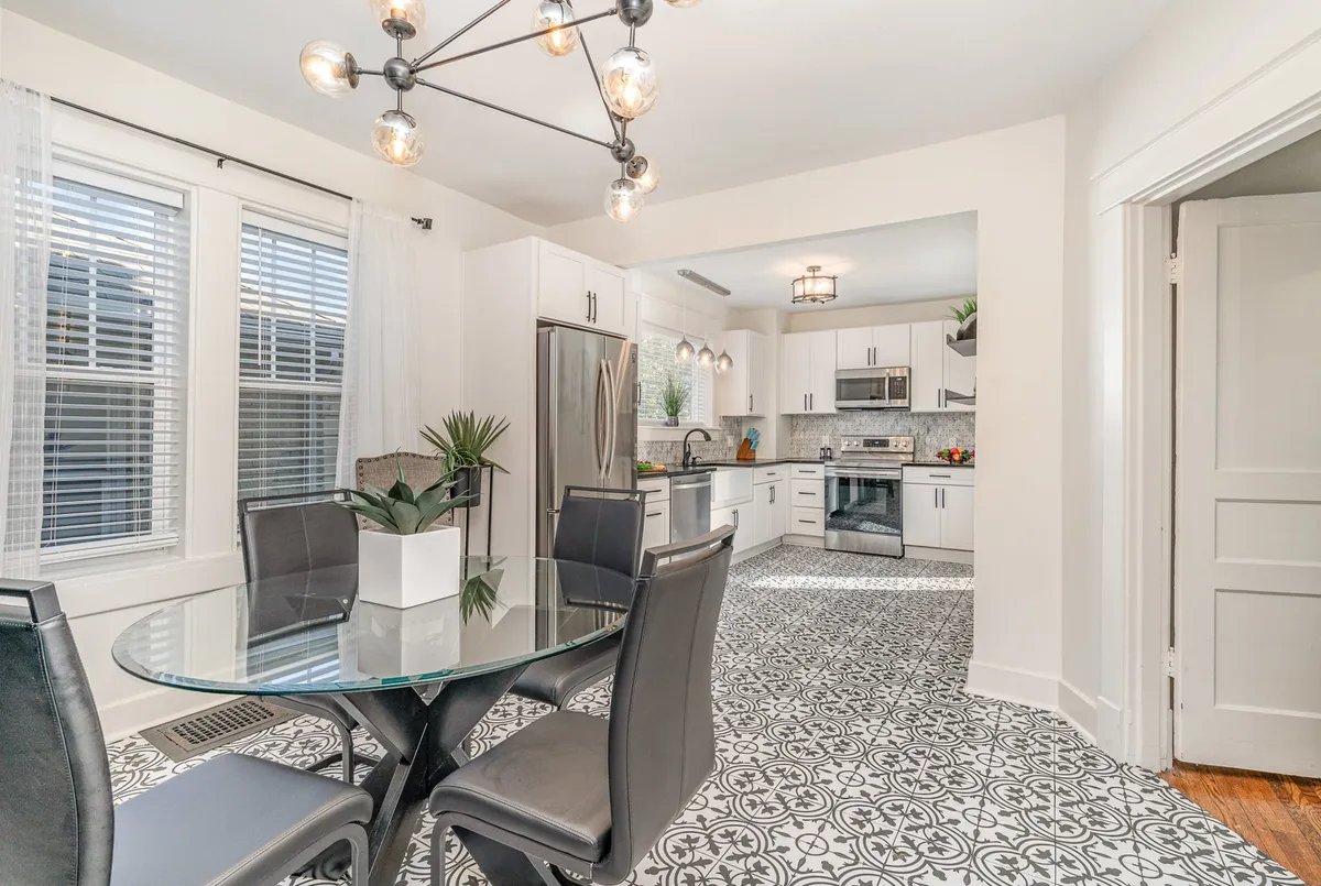 A stylish, open dining area featuring a glass dining table, elegant chandelier, and modern kitchen appliances. Decorative plants add a homey touch to the space, which is clearly designed for family meals and entertaining.