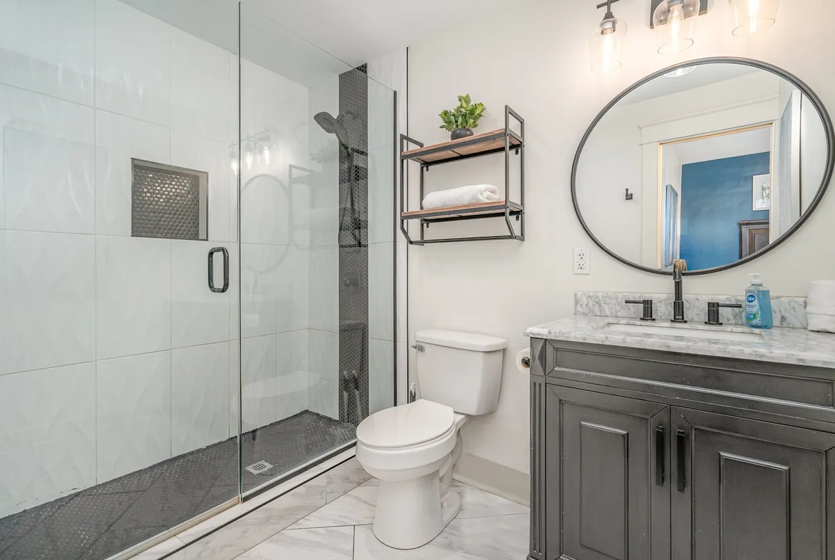A modern, refreshing bathroom with a glass shower enclosure, round mirror, marble countertop, and sleek black shelving. Abundant natural light creates a spa-like atmosphere, highlighting quality amenities and attention to detail.