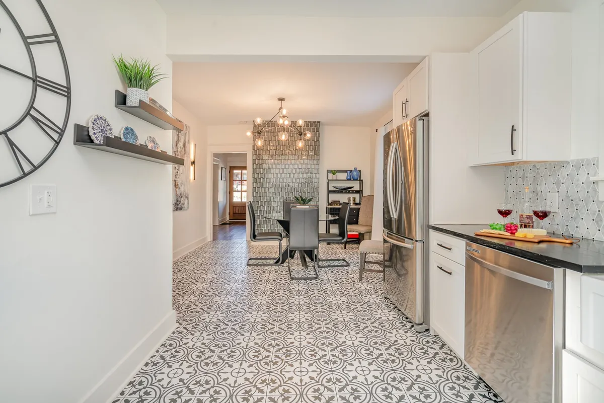 A stylish, modern kitchen and dining area with stainless steel appliances, contemporary furniture, geometric tile flooring, and a decorative clock. The space is both functional and welcoming, ideal for preparing meals or gathering with family.