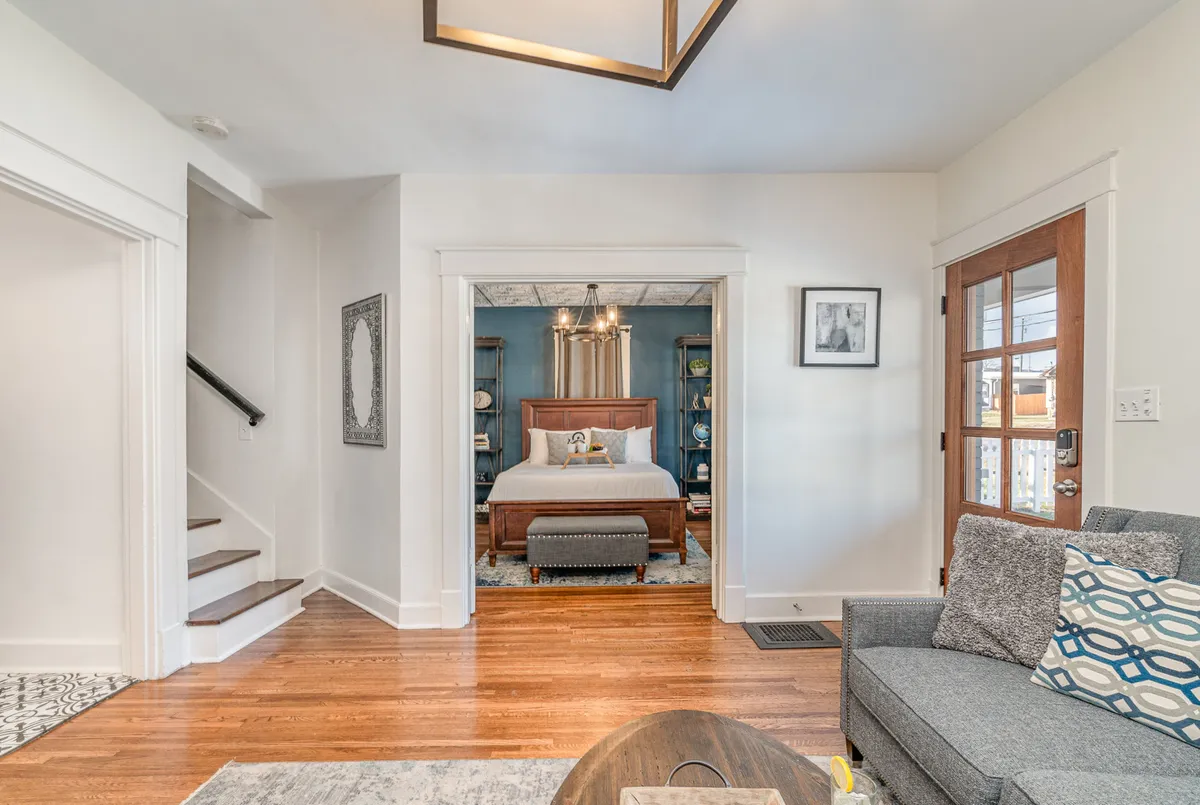 A warm, inviting living area featuring a plush sofa, decorative mirror, and a bedroom visible through the doorway. Natural light streams in, illuminating the cozy space perfect for relaxing or entertaining guests.