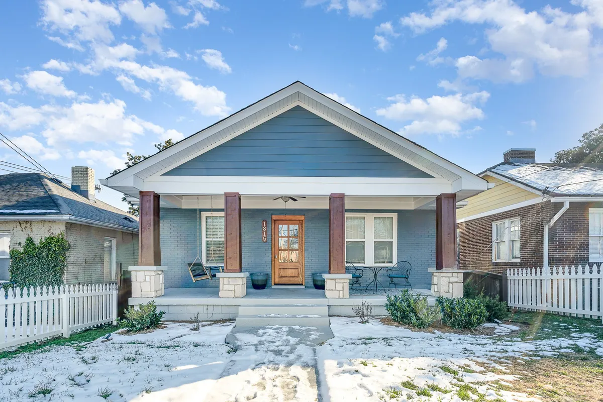 The image showcases the inviting front exterior of the Blue Pearl, with a welcoming front porch featuring classic rocking chairs, a snow-dusted lawn, and a charming white picket fence. The entrance is warm and inviting, hinting at cozy moments and relaxation amid a picturesque winter setting.